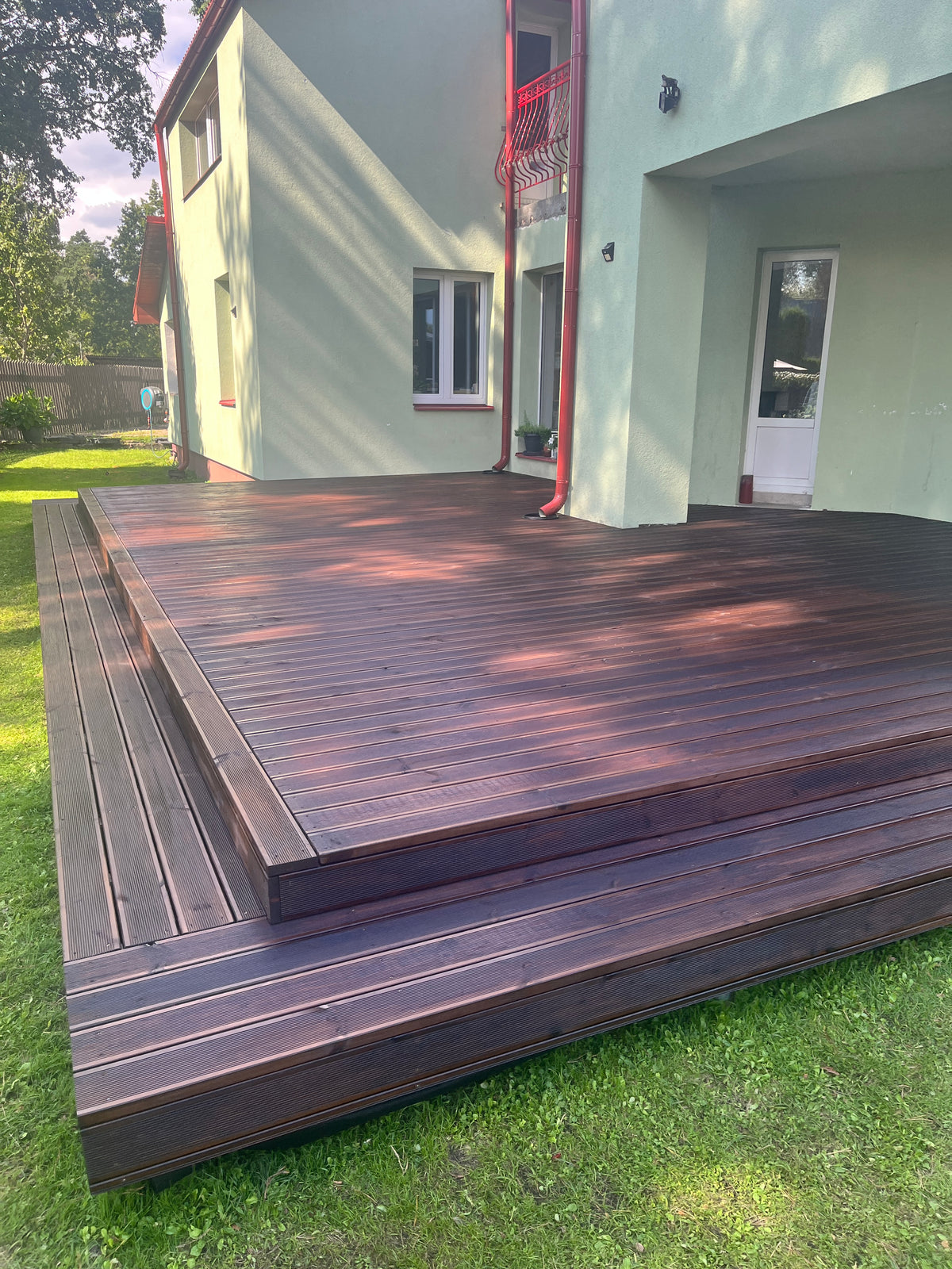 A dark brown wooden outdoor staircase and deck system attached to a light green house, set on a grassy lawn.