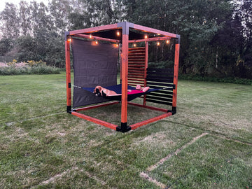 Person lying on a hammock inside a red metal frame structure in a grassy outdoor area.