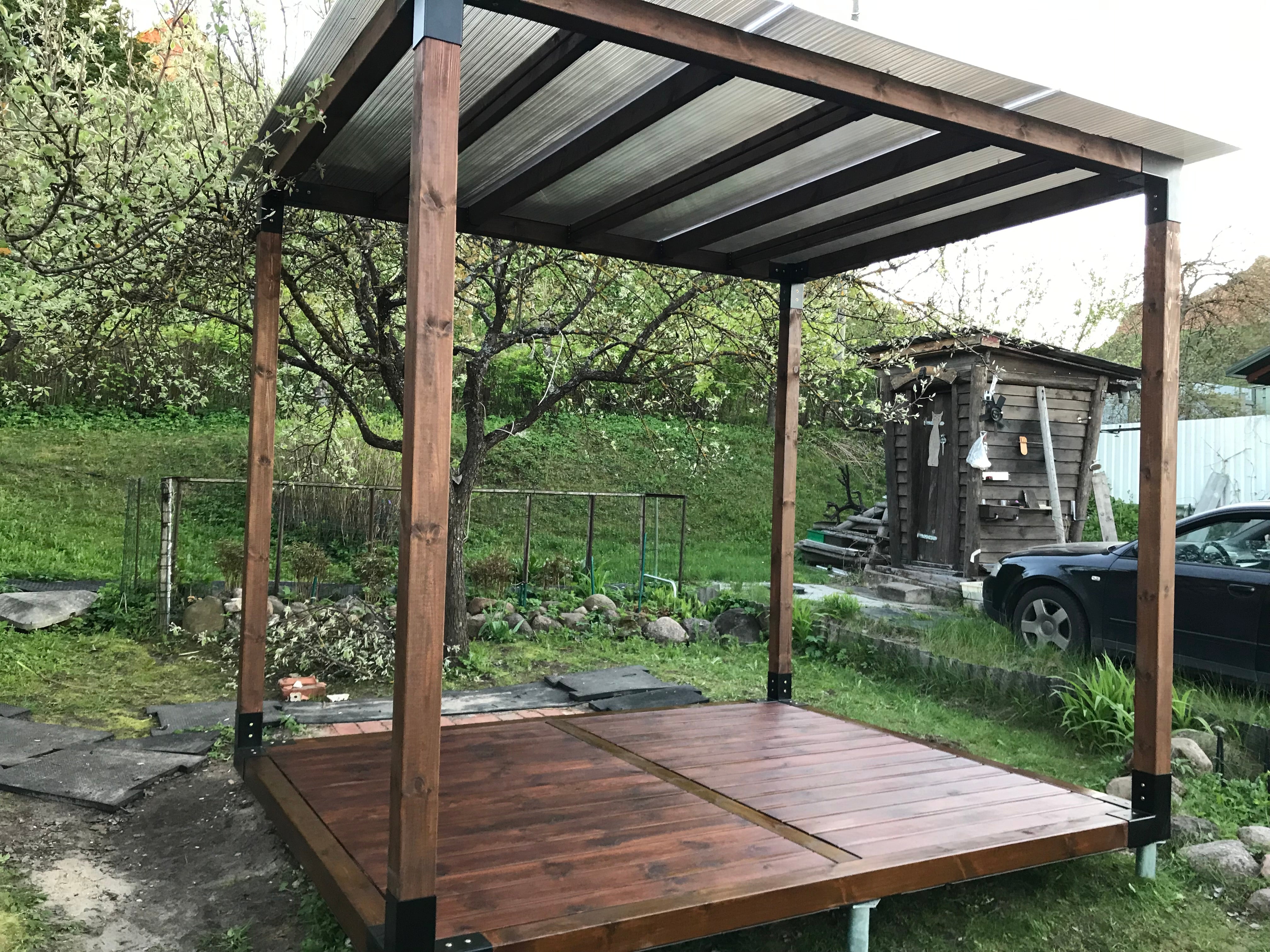 Wooden pergola structure in a garden setting with trees and a car in the background.
