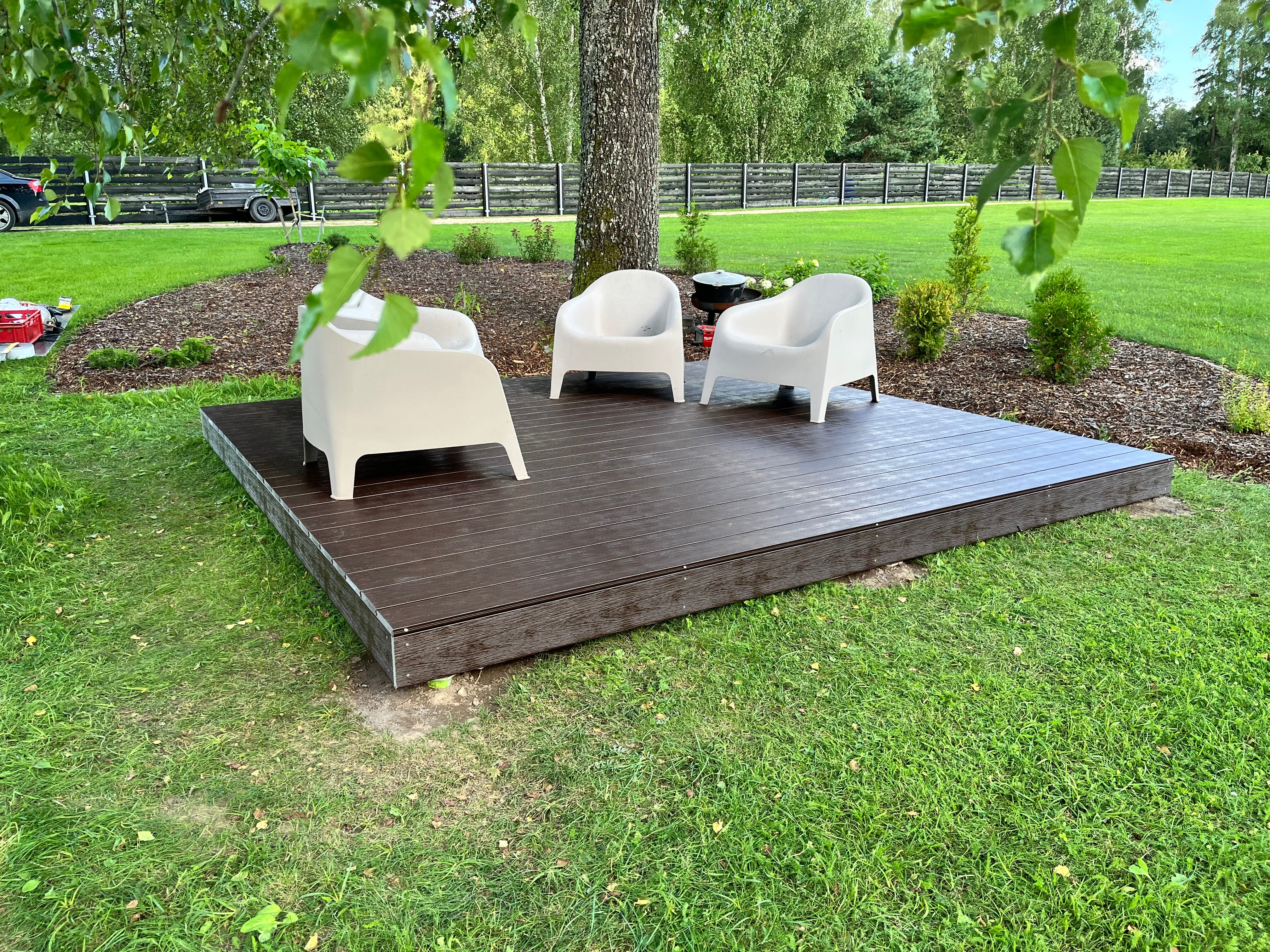 Three white chairs on a wooden platform in a garden setting with green grass and trees.