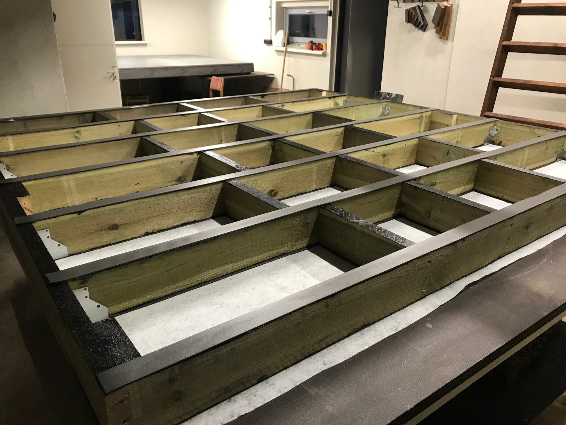 Wooden floor joists on a concrete floor with a kitchen in the background