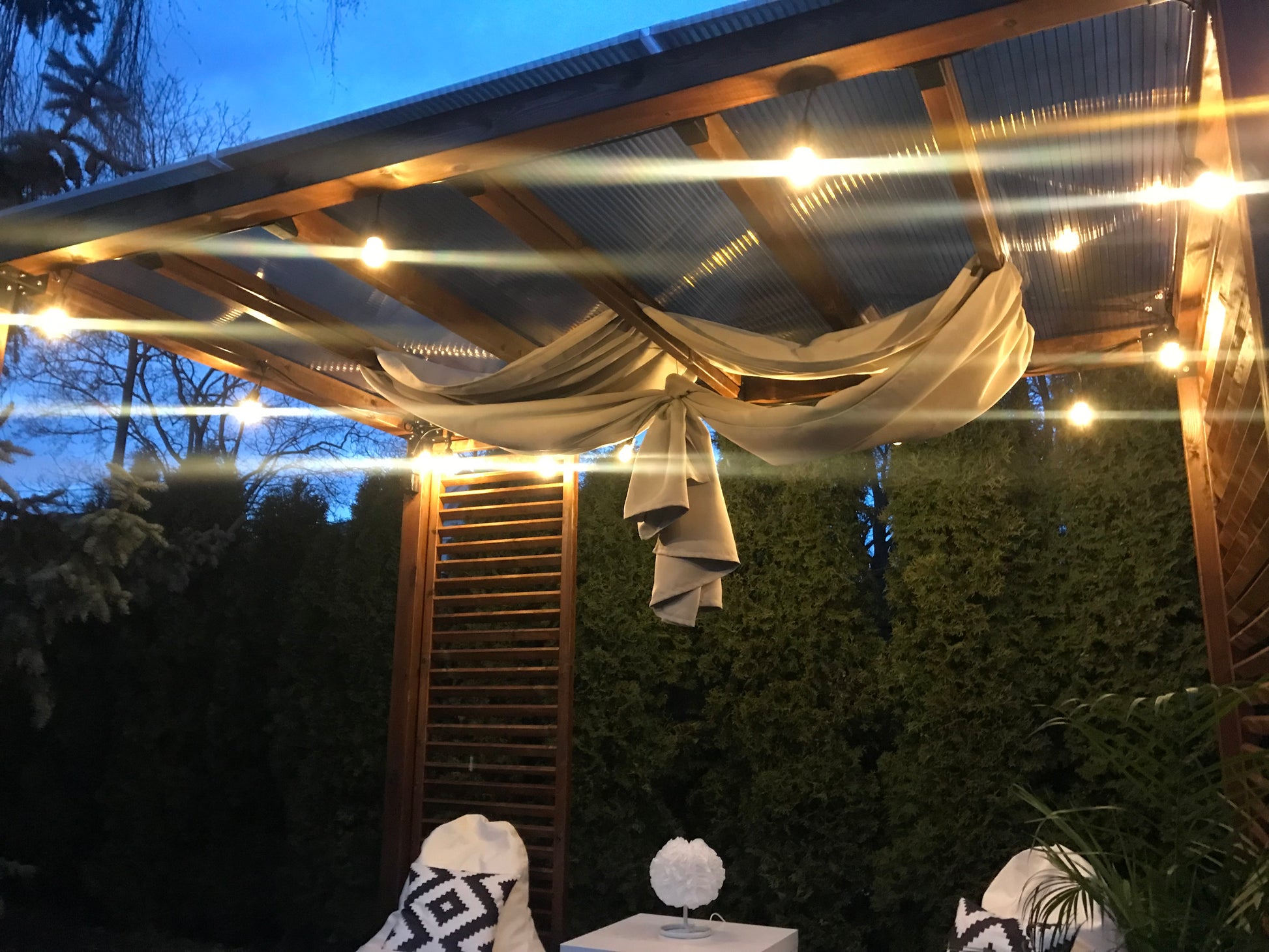 Outdoor setting with string lights and draped fabric on a wooden pergola at dusk.